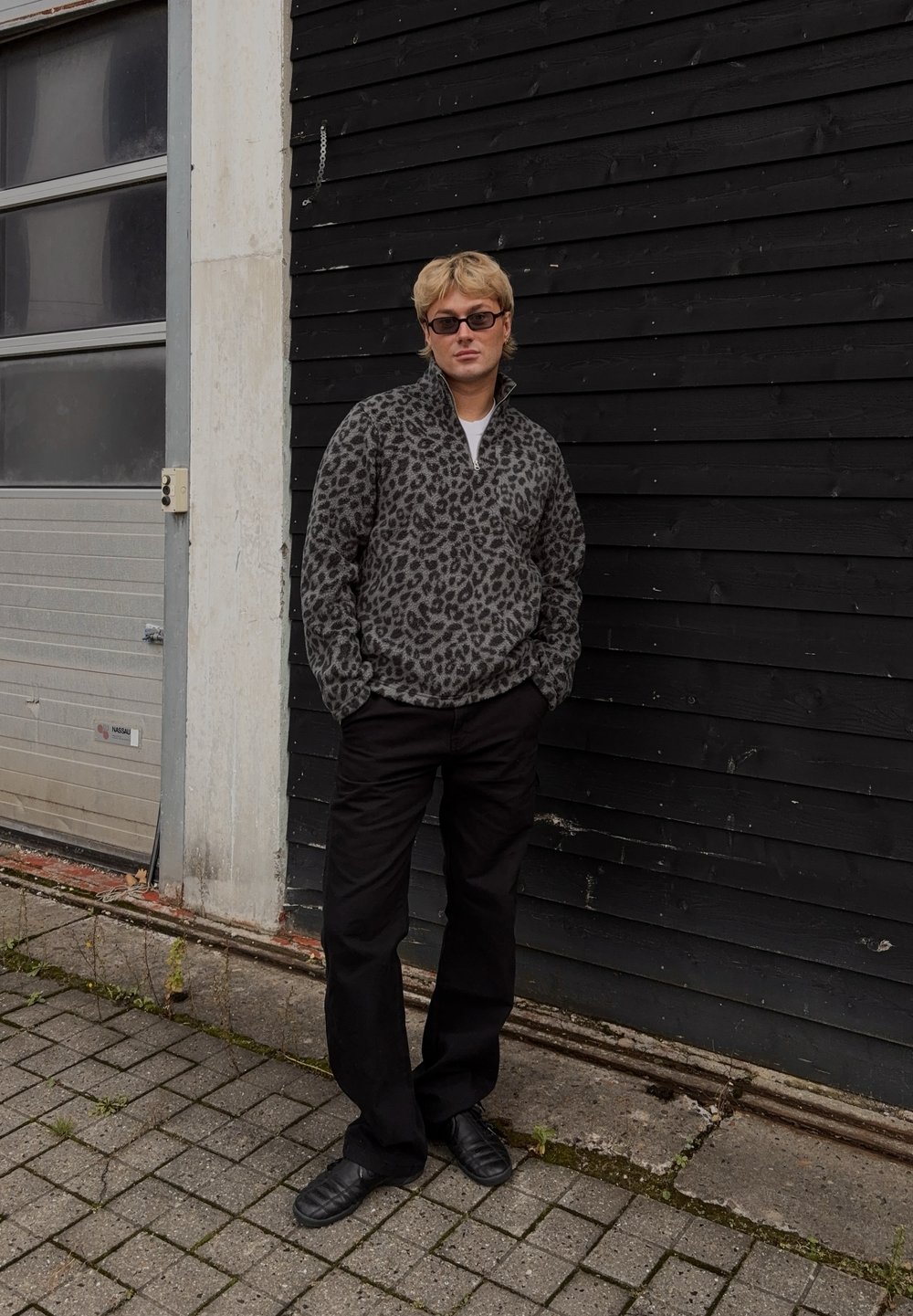 Pulover s polovičnim zadrgo iz flisa z leopardjim vzorcem v sivi in črni barvi, v kombinaciji z ohlapnimi črnimi hlačami in črnimi čevlji brez vezalk, stojijo ob črni steni.