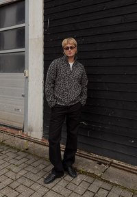 Pulover s polovičnim zadrgo iz flisa z leopardjim vzorcem v sivi in črni barvi, v kombinaciji z ohlapnimi črnimi hlačami in črnimi čevlji brez vezalk, stojijo ob črni steni.