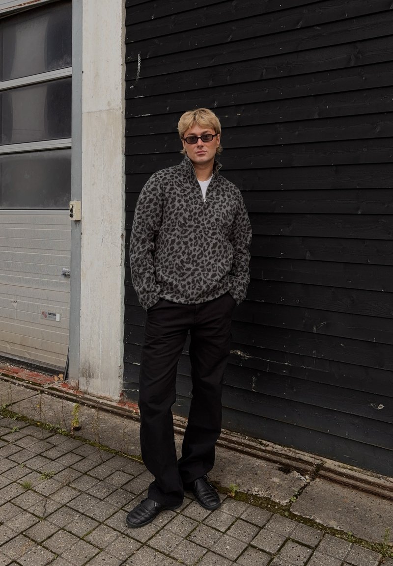 Leopardmønster fleece halv-ble overdel i grå og sort, kombinert med svarte, løse bukser og sorte slip-on sko, står foran en svart vegg.
