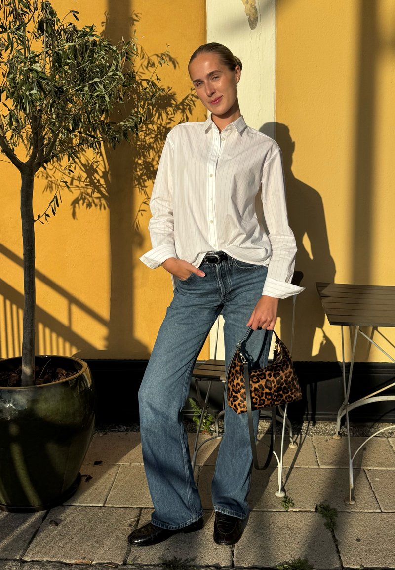 Camicia bianca a righe con bottoni, jeans blu e una borsa leopardata in un ambiente esterno soleggiato con ulivo e muro giallo.