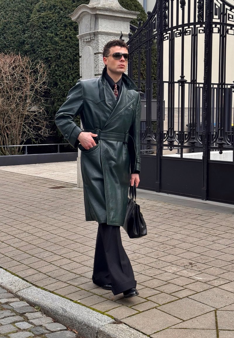 Man loopt op het voetpad en draagt een donkergroene leren trenchcoat, zwarte broek, zonnebril en draagt een zwarte handtas nabij een gesloten toegang.