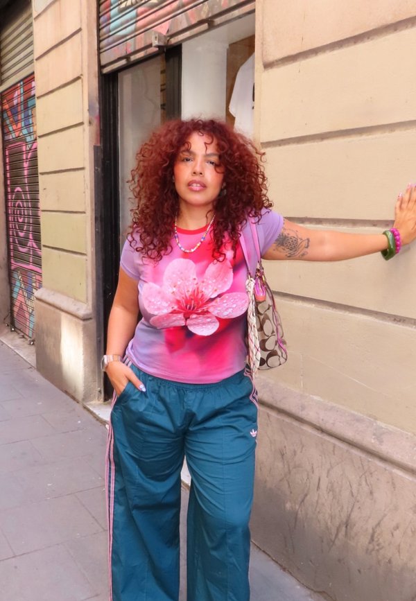 Mujer con cabello rizado rojo que lleva una camiseta rosa con estampado de flores y pantalones azules, de pie contra la pared beige de un edificio en una acera de la ciudad.