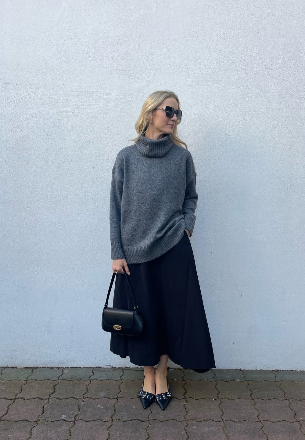 Pelēka turtlenecka džemperis, melna plūstoša svārku kleita, melna rokassoma un strupes kurpes ar rotājumiem. Modelis stāv pret gaismas sienu.