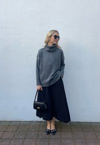 Pelēka turtlenecka džemperis, melna plūstoša svārku kleita, melna rokassoma un strupes kurpes ar rotājumiem. Modelis stāv pret gaismas sienu.