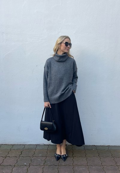 Suéter de cuello alto gris, falda negra fluida, bolso negro y tacones de punta con adornos. La modelo está de pie contra una pared clara.
