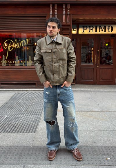 Hombre con cabello rizado, que lleva una chaqueta marrón y vaqueros rotos, está de pie en la acera frente al restaurante mexicano Primo con fachada de madera.