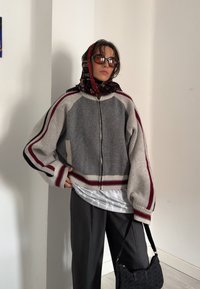 Casaco cinzento de malha com riscas vermelhas e azul marinho, com fecho de zip, mangas oversized, combinado com um lenço estampado e uma mala preta.