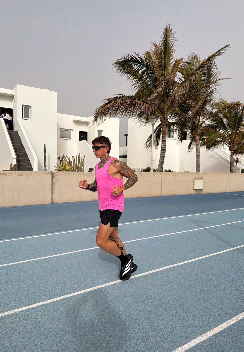 Homme avec des tatouages et des lunettes de soleil faisant du jogging sur une piste extérieure bleue près de bâtiments blancs et de palmiers sous un ciel clair.