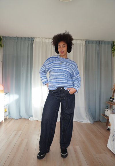 Suéter de punto a rayas azules y blancas combinado con vaqueros anchos oscuros y zapatos negros. La modelo está de pie en una habitación luminosa con cortinas suaves.