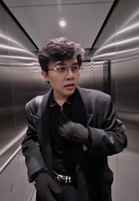 Jeune personne portant des lunettes et un manteau noir ajuste son col, debout à l'intérieur d'un ascenseur en acier inoxydable aux parois réfléchissantes.