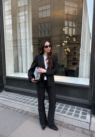 Blazer y pantalones negros a medida sobre una camisa blanca. Bolso negro con un brillo metálico. Gafas de sol cuadradas. Fondo de ventana reflectante.