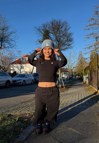 Joven mujer con un gorro gris de LA, top corto negro y pantalones anchos, sonriente, de pie en una acera soleada en un barrio suburbano con coches estacionados cerca.
