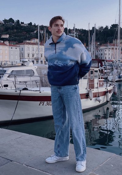 Sudadera de fleece en azul y blanco con estampado de montañas y nubes, con cuello de media cremallera; combinada con jeans de mezclilla ligeros y sueltos de color azul claro y zapatillas blancas.