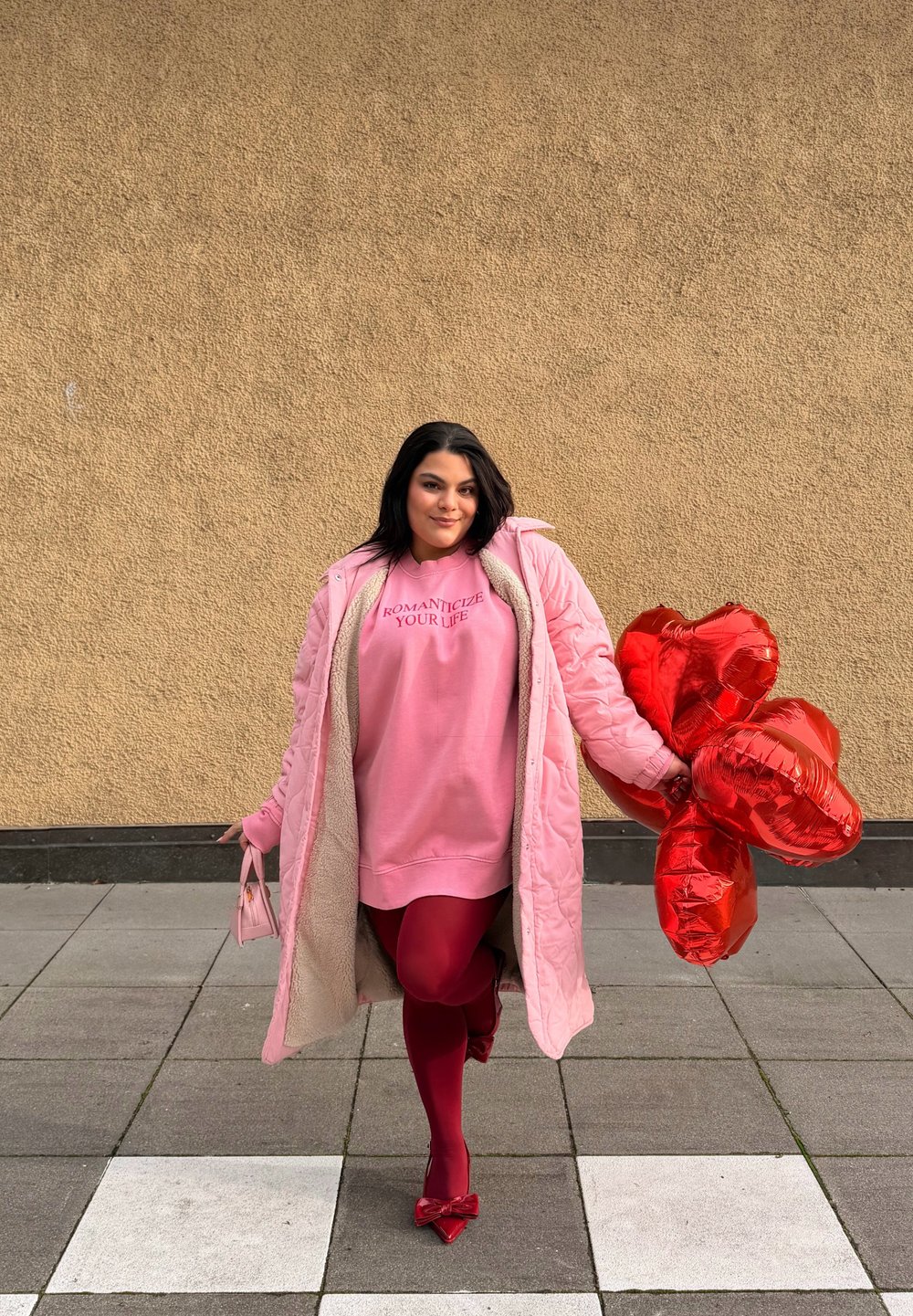Femme en manteau rose et sweat-shirt tenant des ballons rouges en forme de cœur et un petit sac à main rose, debout sur un trottoir carrelé devant un mur beige.