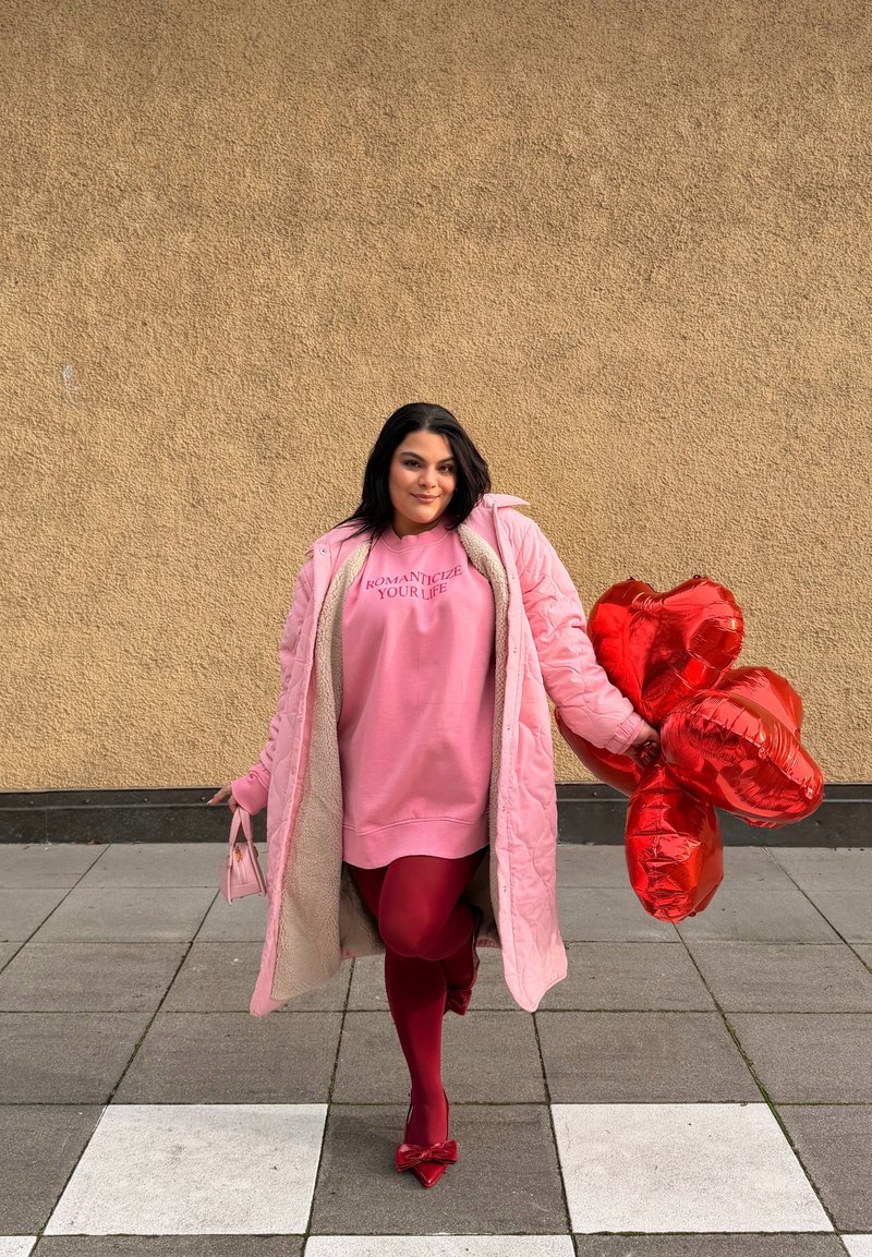 Femme en manteau rose et sweat-shirt tenant des ballons rouges en forme de cœur et un petit sac à main rose, debout sur un sol carrelé devant un mur beige.