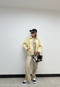 Κίτρινο υφασμάτινο cardigan πάνω από μπεζ μπλούζα, σε συνδυασμό με ανοιχτόχρωμο παντελόνι και μαύρα αθλητικά παπούτσια. Αξεσουάρ περιλαμβάνουν ένα μαύρο καπέλο και μια τσάντα με σχέδιο.