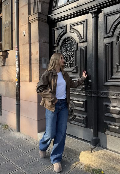 Chaqueta de cuero marrón, camiseta blanca, vaqueros de pierna ancha azules y botas marrones. De pie junto a una puerta negra ornamentada con detalles intrincados.