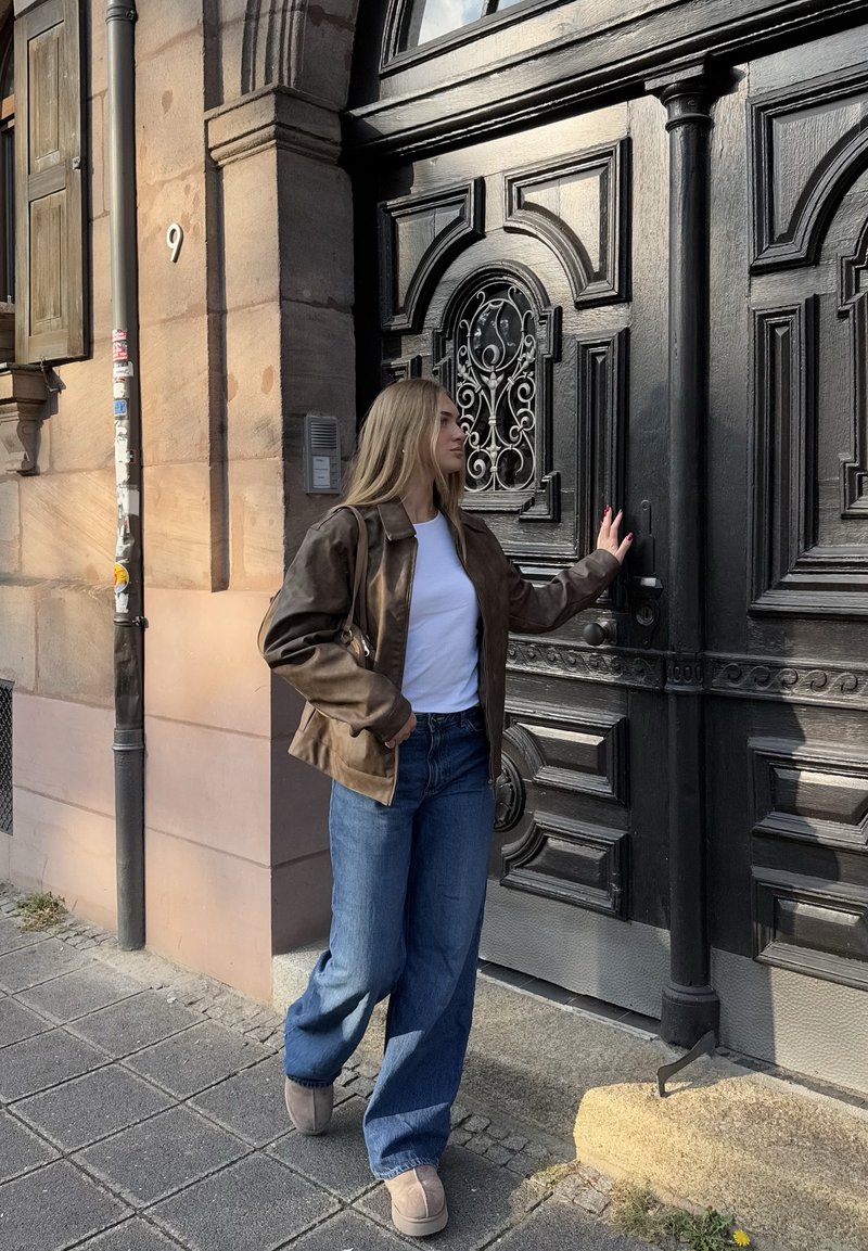 Jaqueta de couro castanha, t-shirt branca, jeans azul de perna larga e botas beijes. De pé ao lado de uma porta negra ornamentada com detalhes intrincados.