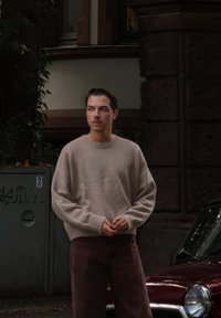 Beige strikket sweater og brune fløjlsbukser. Står nær en grå elskab med en mørkerød bil delvist synlig i baggrunden.