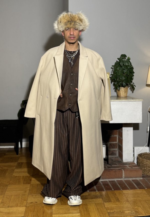 Uomo con grande cappotto beige sopra un gilet di pelle marrone, pantaloni a righe marroni, sneakers bianche e cappello peloso, in piedi all'interno vicino a un camino.