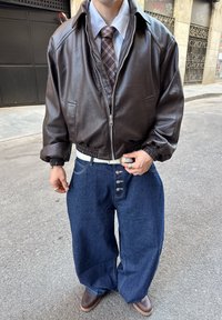 Brązowa skórzana kurtka typu bomber z elastycznymi mankietami, jasnoniebieska koszula z kołnierzykiem, wzorzysty krawat, luźne niebieskie dżinsy, brązowe buty.