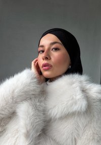 Abrigo de piel sintética blanca con textura esponjosa, usado sobre un hijab negro. El abrigo tiene un cuello voluminoso y un estilo elegante, sobre un fondo neutro.