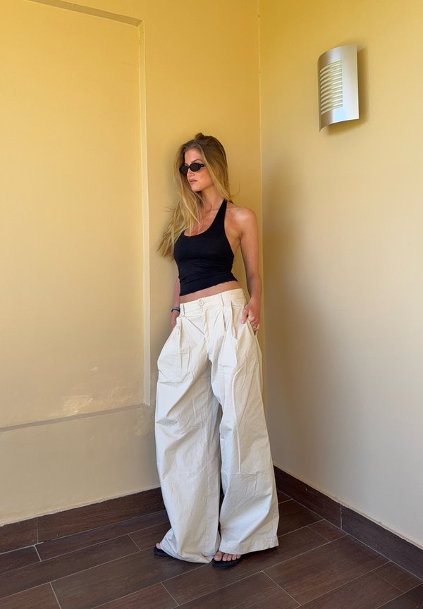 Vrouw met lang blond haar, gekleed in een zwart tanktop, brede witte broek, zwarte sandalen en ovale zonnebril, staand tegen een beige muurhoek.