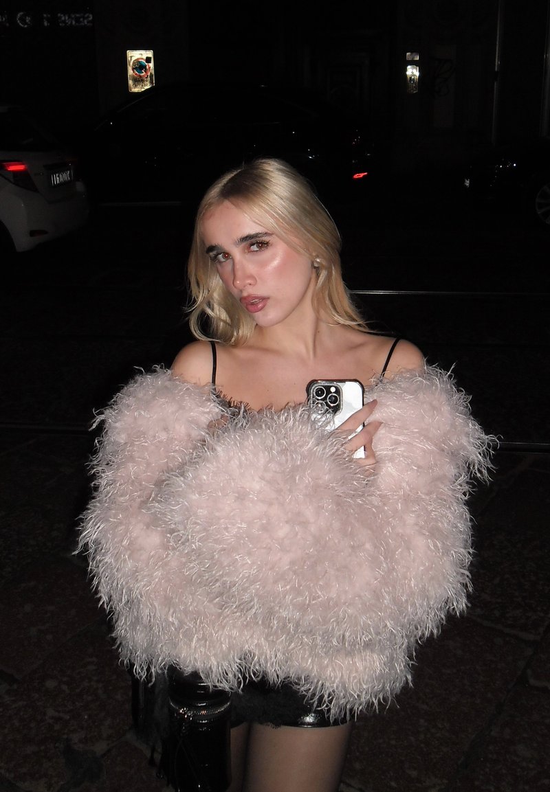 Vêtement d'extérieur rose duveteux avec une finition texturée, porté par-dessus une tenue noire. La personne tient un téléphone, avec un arrière-plan sombre derrière elle.