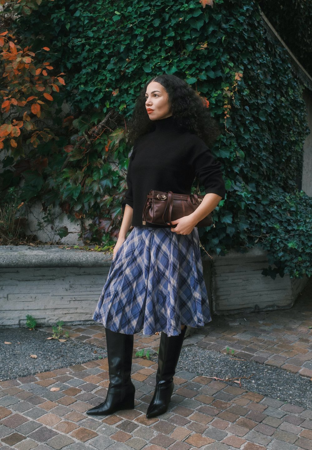 Uma mulher veste um suéter de gola alta preto, uma saia xadrez azul e botas altas pretas, segurando uma mala castanha diante de um fundo de hera verde.