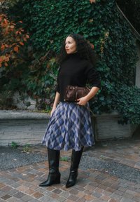 Uma mulher veste um suéter de gola alta preto, uma saia xadrez azul e botas altas pretas, segurando uma mala castanha diante de um fundo de hera verde.