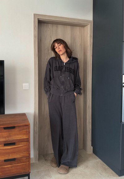Mujer con cabello castaño, vestida con una sudadera con capucha gris oscuro y pantalones anchos, de pie de forma relajada en un marco de puerta dentro de una habitación moderna.