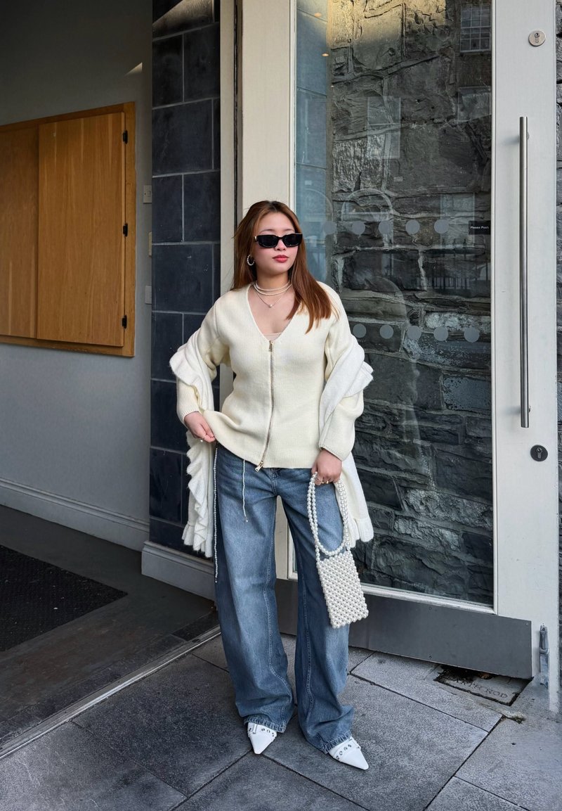 Femme en pull à fermeture éclair blanc, jeans à jambes larges, chaussures blanches à bout pointu, lunettes de soleil, tenant un sac à main en perles, debout près d'une porte en verre avec un mur en pierre en arrière-plan.