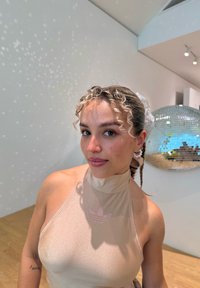 Top bege, sem mangas, com um gola alta e um leve brilho; apresenta um logo Adidas em rosa claro; cabelo encaracolado; o fundo inclui uma bola de discoteca refletiva.