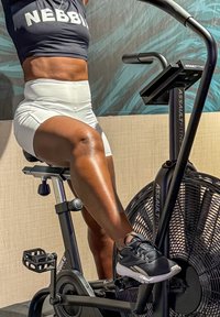 Svart Assault Fitness-sykkel med vifte, utstyrt med teksturerte pedaler og justerbart sete, okkupert av en person iført svart topp og hvite shorts.