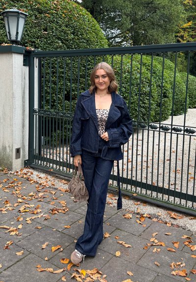 Chaqueta de mezclilla y pantalones acampanados en azul oscuro, combinados con una blusa de estampado de leopardo. Llevando un pequeño bolso beige, de pie en un camino con hojas de otoño.