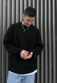 Jeune homme portant un pull en maille noir et un jean bleu clair, debout devant des lattes de bois grises verticales, ajustant sa manche et regardant vers le bas.