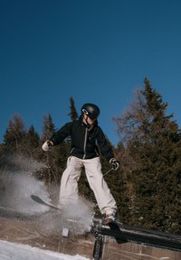 Skieur en veste noire et pantalon blanc glissant sur une rampe avec ses bâtons, neige éclaboussant, avec des arbres à feuilles persistantes et un ciel bleu clair en arrière-plan.