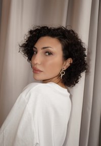 Žena s kučeravými tmavými vlasmi v bielom tričku a zlatými kruhovými náušnicami sa pozerá cez plece na pozadí béžovej záclony.