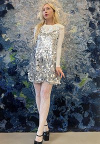 Robe en sequins argentés avec un col floral texturé, portée sur des collants en dentelle blanche et des chaussures à plateforme noires, se détachant sur un fond bleu texturé.