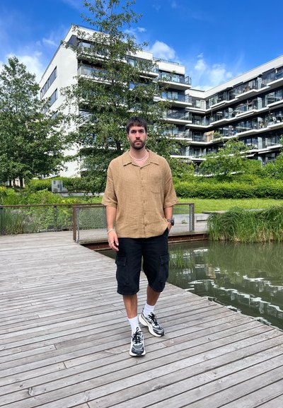 El atuendo informal incluye una camisa de cuadros marrón de manga corta, pantalones cortos cargo negros y zapatillas en blanco y negro, con una pasarela de madera y vegetación de fondo.