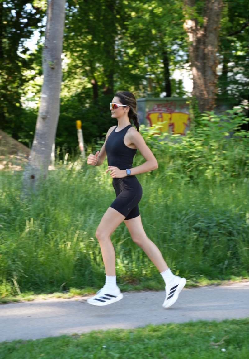 Mladá žena v černém sportovním oblečení a slunečních brýlích běží po parkové cestě obklopené zelenými stromy a trávou.