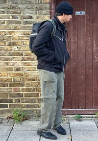 Casaco preto com fecho éclair, calças cargo verdes-oliva com bolsos laterais, sapatos pretos e uma mochila preta com alças verdes. Fundo de parede de tijolo.