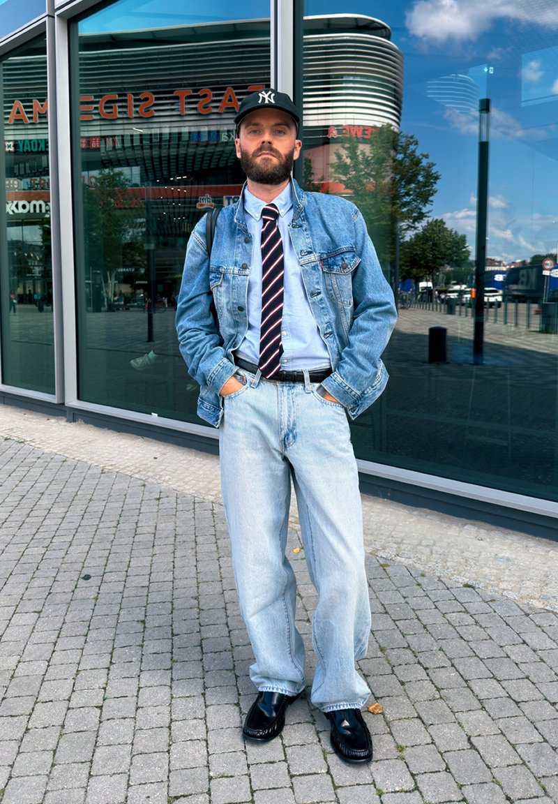 Homme barbu portant une veste en jean et un jean, une cravate rayée, une casquette noire et des chaussures habillées, debout sur le trottoir de la ville avec un immeuble en verre réfléchissant derrière lui.