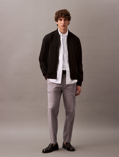 Chaqueta bomber negra sobre una camisa blanca, combinada con pantalones gris claro y zapatos de cuero oscuros. Texturas suaves y corte entallado.