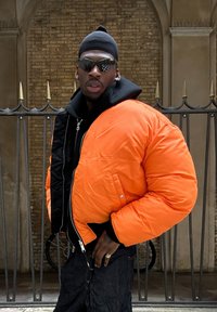 Veste rembourrée orange vif avec une capuche noire, dotée d'une fermeture éclair et d'accents aux épaules. Portée sur un haut noir, présentée devant un mur en briques.