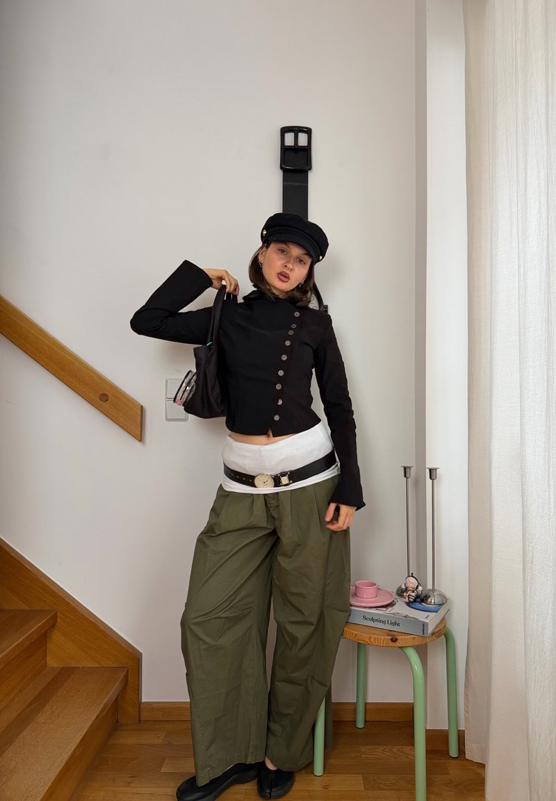 Joven mujer con sombrero negro y top, pantalones verdes holgados con un cinturón ancho, de pie en el interior cerca de unas escaleras de madera y una pequeña mesa con libros y velas.