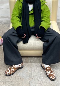 Καφέ και λευκά παπούτσια με σχέδιο και επίπεδη σόλα, σε συνδυασμό με σκούρους φαρδείς παντελόνες και πράσινο φλις μπουφάν, καθισμένος σε ανοιχτόχρωμη καρέκλα.