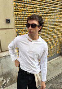 Chemise blanche à manches longues avec un découpage, jean noir avec une ceinture et lunettes de soleil. L'arrière-plan présente une surface à motifs gris et jaune.