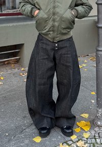 Pantalon en denim foncé ample à taille haute, avec de grandes jambes larges et une fermeture à trois boutons. Associé à une veste bomber verte.