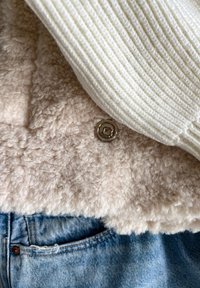 Beige falsk pelsjakke med en cremet ribbet krave og guldtrykknap. Blå denimjeans synlige nederst, der viser tekstureret stof.